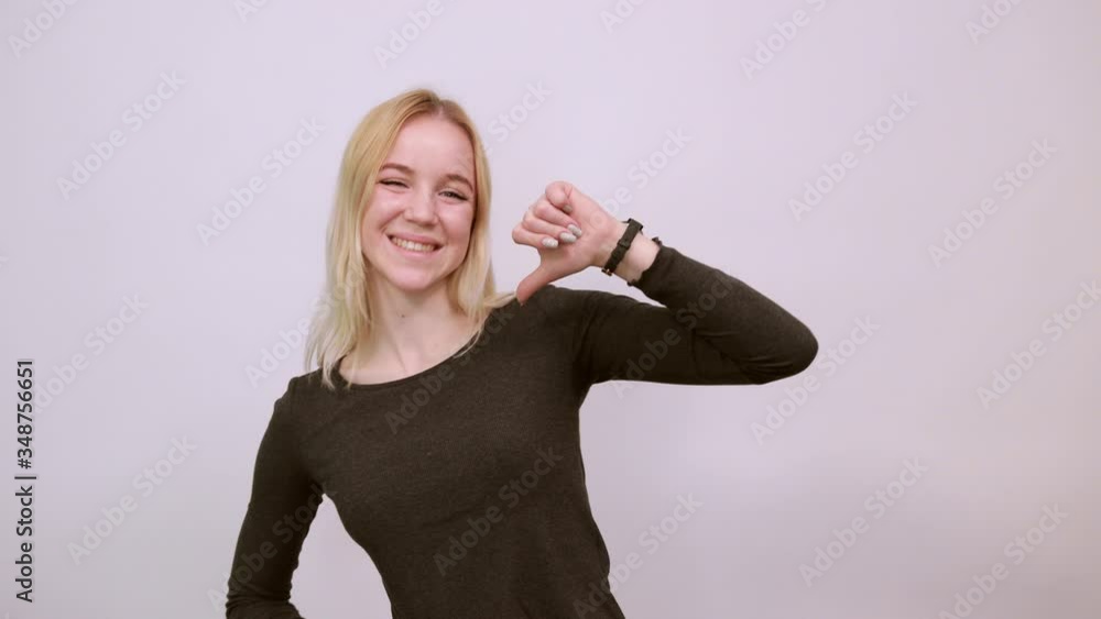Young Blonde Woman In Black Sweater With Stylish Watch On White Background, Happy Girl Smiles, Shows Her Thumb Finger. The Concept Of Successful And Confident People Young Blonde Woman In Black Sweater With Stylish Watch On White Background, Happy Girl Smiles, Shows Her Thumb Finger. The Concept Of Successful And Confident People