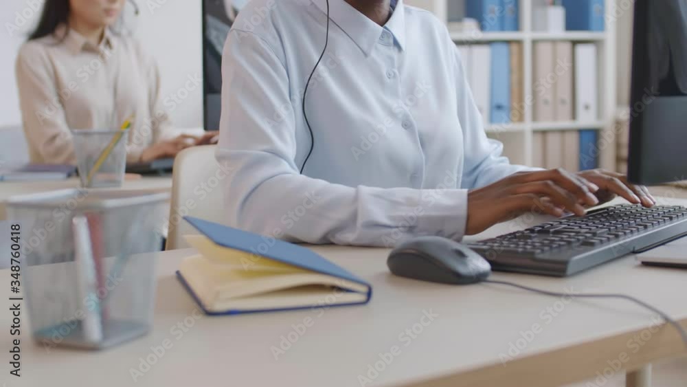 Lockdown of unrecognizable female African office worker wearing light blue shirt sitting at workplace and typing on computer keyboard while her colleague working on background Lockdown of unrecognizable female African office worker wearing light blue shirt sitting at workplace and typing on computer keyboard while her colleague working on background