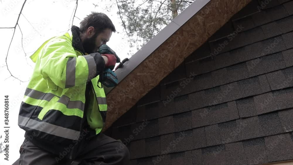 Young man with safety yellow jacket stands on ladder next to the sheds roof and screwing plate to wooden log with a cordless screwgun. One person. Roofer. frog perspective. Young man with safety yellow jacket stands on ladder next to the sheds roof and screwing plate to wooden log with a cordless screwgun. One person. Roofer. frog perspective.