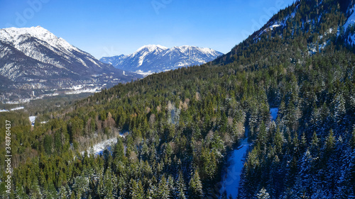 The mountainous Alpine landscape. High snow-capped mountains with sharp peaks, rocks and cliffs under a clear blue sky. Large coniferous forest at the foot of the mountains. Winter season of skiing. 