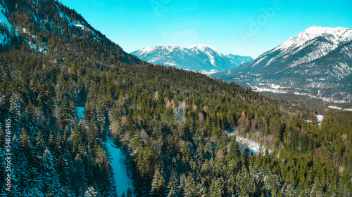 The mountainous Alpine landscape. High snow-capped mountains with sharp peaks, rocks and cliffs under a clear blue sky. Large coniferous forest at the foot of the mountains. Winter season of skiing. 