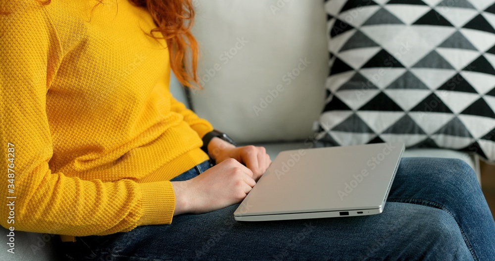 Naklejka premium Close up of redhead girl using silver laptop while sitting on sofa in living room at home.