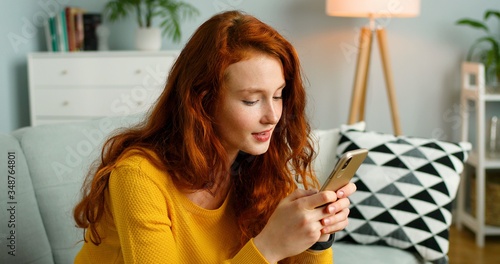 Schilderij op canvas Portrait of beautiful cheerful redhead girl relax sit on sofa at home using cellphone