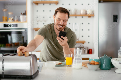 Handsome man preparing breakfast at home. Young man using the phone in kitchen.