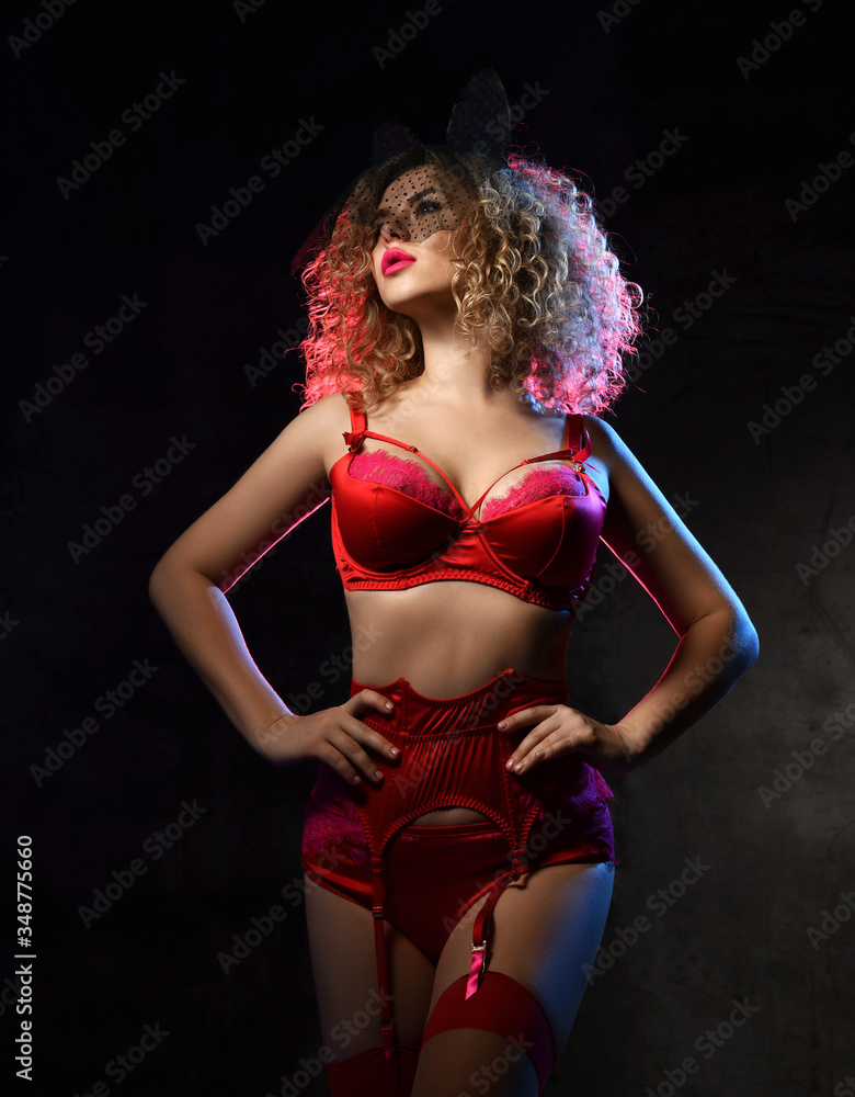 Obraz premium Woman with blonde curly hair in red lace sexy lingerie, garter belt, stockings and veil stands with her hands on waist