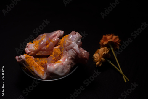 A bowl of raw chicken pieces sprinkled with turmeric powder decorated with dried flowers in a dark copy space background. Food and product photography.