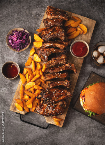 Grilled pork ribs and fried potato wedges on a wooden board, burger and cola glass, sause. Top view, fast food.