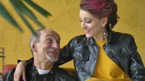 Emotional family portrait of adult daughter and senior father in loft room with houseplants. Laughing man and girl in black leather jackets in punk style on yellow wall background