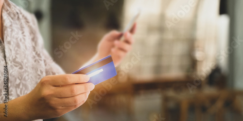 Cropped image of customer's hand holding a smartphone and credit card for doing a payment by using a credit card over blurred restaurant as background.