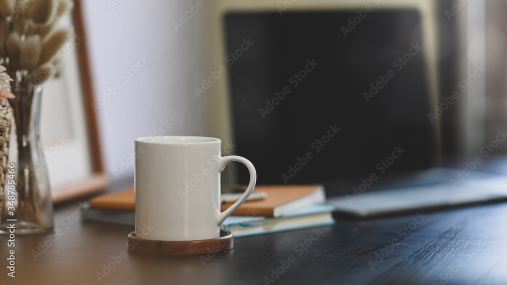 Coffee cup putting on black working desk that surrounded by stack of notebook, wild grass in vase, empty picture frame and computer laptop. Coffee cup putting on black working desk that surrounded by stack of notebook, wild grass in vase, empty picture frame and computer laptop.