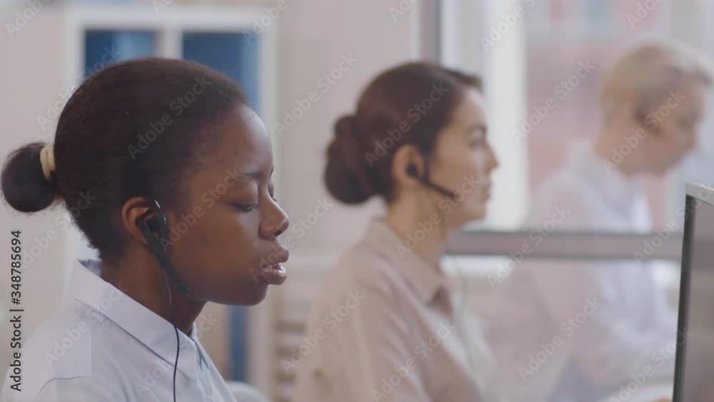 Close up of African woman wearing blouse and headset giving advice to interlocutor by phone and two diverse women working on background Close up of African woman wearing blouse and headset giving advice to interlocutor by phone and two diverse women working on background