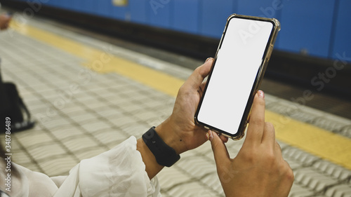 Fototapeta Naklejka Na Ścianę i Meble -  Cropped image of attractive woman's hands holding a black smartphone with white blank screen over empty railway platform as background.