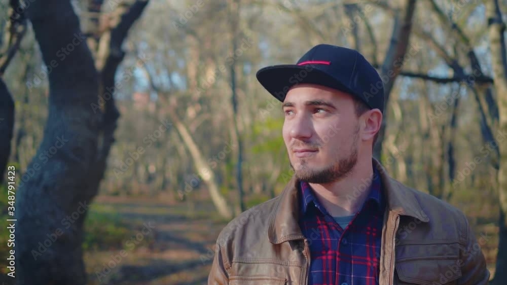 Young man in the autumn park. The guy in the leather jacket and baseball cap looks away. The guy on the background of trees without foliage. Young man in the autumn park. The guy in the leather jacket and baseball cap looks away. The guy on the background of trees without foliage.