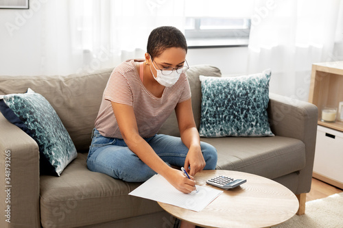accounting, taxes and finances concept - young african american woman in glasses with papers and calculator wearing face protective medical mask for protection from virus disease at home