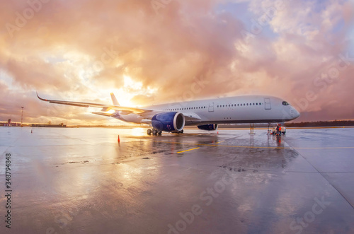 Passenger commercial wide-body aircraft parked with a wet apron at the airport during evening rain at sunset with beautiful sky.