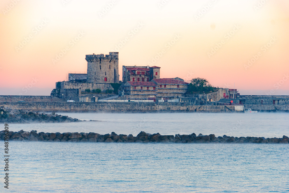 view of the medieval building tower in Socoa, Saint Jean de Luz, Phrase, at dawn, under the golden light of the sun, from the other side of the bay, behind the rocks and the sea ...