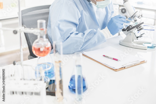 A scientist wearing a blue protective lab coat  glove  and mask looking into a microscope in a laboratory setting with test tubes.