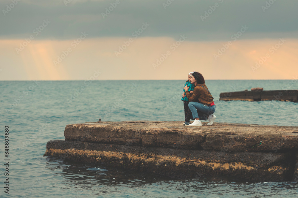 Mother and daughter stand on a breakwater by the sea and look at the horizon at sunset.