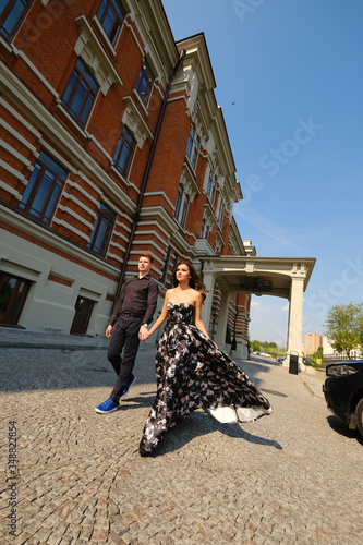 Young beautiful couple - a girl in a magnificent dress and a guy in a black shirt and trousers walk along the isoic brick building