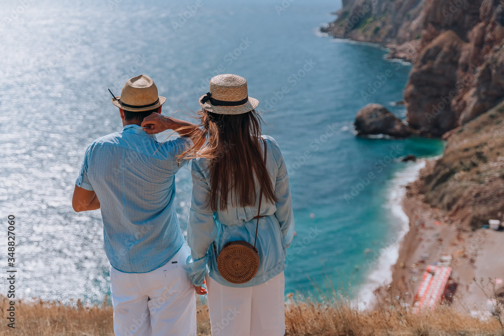 The couple stands with their back to the audience and look down at the beautiful seascape. They are wearing hats, blue shirts and white shorts.
