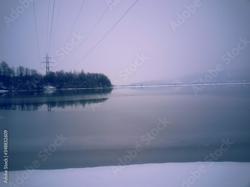 frozen lake in winter season close to the village with forest reflecting in water