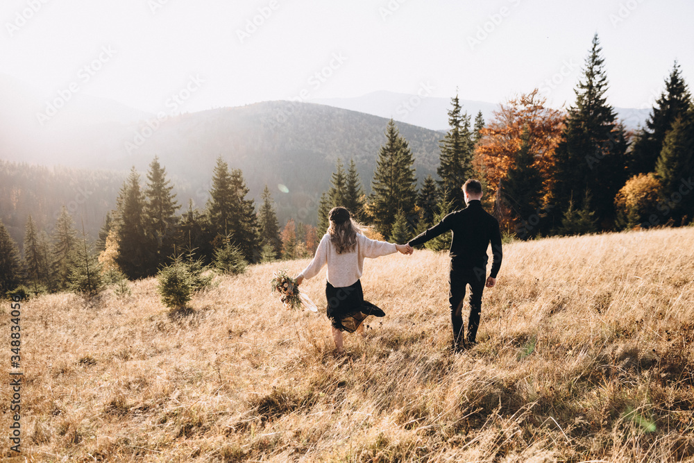 Stylish model couple in the autumn mountains. A young guy and a girl run along the slope against the background of the forest and mountain peaks at sunset. Girl holds in her hands a bouquet of flowers