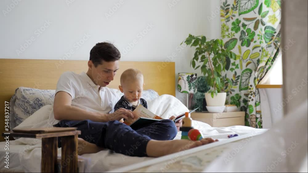 A young father and his baby son read a children's book sitting on a bed at home A young father and his baby son read a children's book sitting on a bed at home
