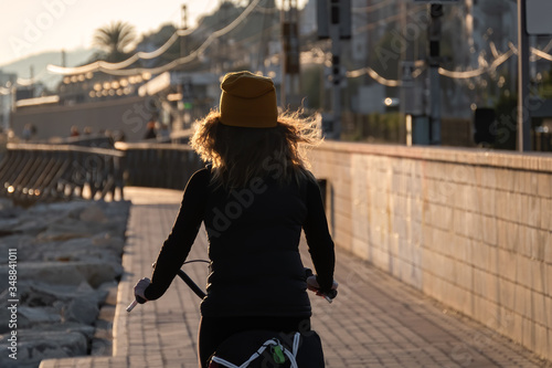 Young girl in a yellow hat with curly hair fluttering in the wind riding a bicycle at sunset on the promenade road of Maresme, Catalonia, Spain. photo