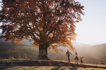 Stylish young family in the autumn mountains. A guy and a girl with their daughter walk together under a large old tree against the background of a forest and mountain peaks at sunset.