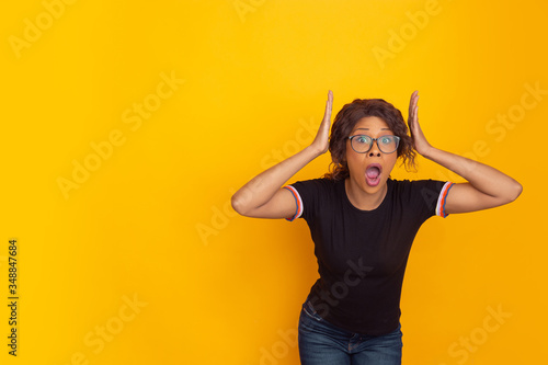 Crazy shocked with head in hands. African-american young woman's portrait isolated on yellow studio background. Beautiful curly model. Concept of human emotions, facial expression, sales, ad, youth.