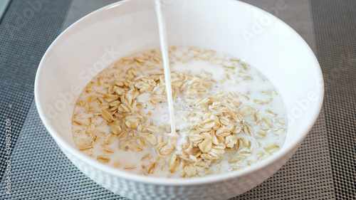 person pours fresh milk into ceramic bowl with tasty porridge cereals on grey table in hotel restaurant extreme close view