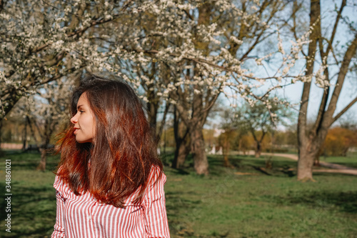 A young girl in a striped shirt with long hair stands against the background of an Apple orchard in early spring, a blooming Apple tree with white flowers.
