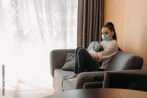 upset young woman in protective mask sitting on sofa and hugging pillow