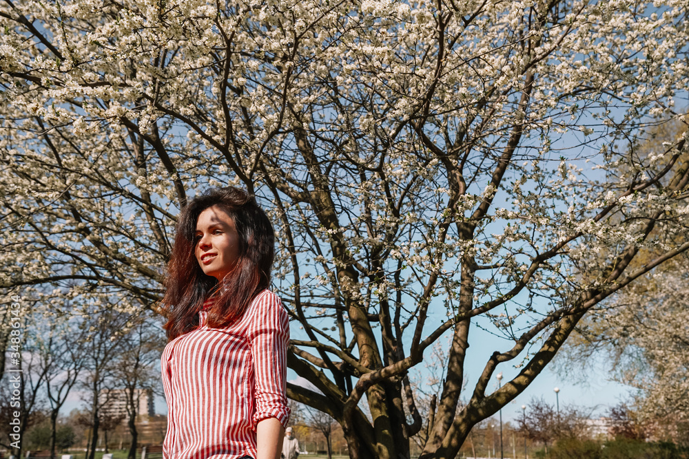 A young girl in a striped shirt with long hair stands against the background of an Apple orchard in early spring, a blooming Apple tree with white flowers. A young girl in a striped shirt with long hair stands against the background of an Apple orchard in early spring, a blooming Apple tree with white flowers.