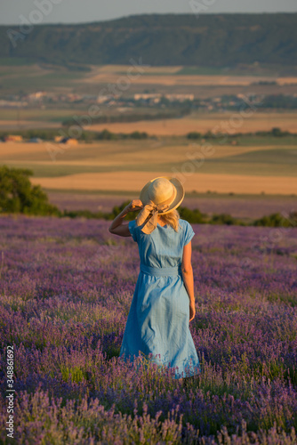 Slender young girl in a simple blue dress and a hat in a lavender field looks into the distance