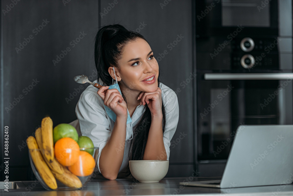 smiling, dreamy woman smiling while holding spoon near laptop, bowl and fruits in kitchen smiling, dreamy woman smiling while holding spoon near laptop, bowl and fruits in kitchen
