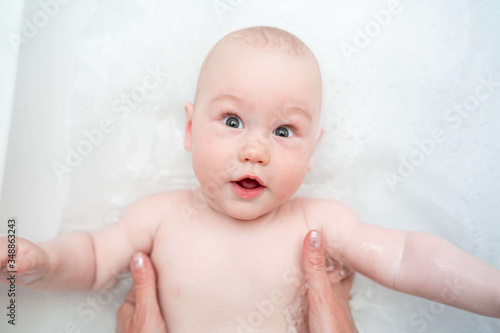 A baby boy bathes in a bath. Caring mother's hands. Happy infant while taking a bath. Care of a newborn baby content. Happy funny baby laughing and bathed in bath. Top view.