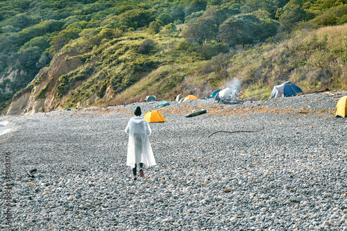young girl in a raincoat walking along a pebble beach towards a beach camping. Tourism and outdoor concept.