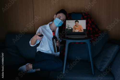 young woman in medical mask holding bottle of wine and tv remote controller while sitting on sofa near laptop with smiling asian boyfriend on screen