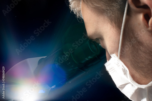 Up-close photo of a white male scientist wearing face maskusing a binocular microscope against a black background. Flash used for effect.