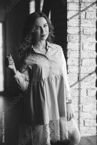 Beautiful young woman with curly dark hair and red lips in a powdery dress is standing near the window in the loft studio. Soft selective focus. Black and white art photo.
