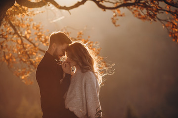 Stylish young couple in the autumn mountains. A guy and a girl hug together under a large old tree on a background of a forest and mountain peaks at sunset.