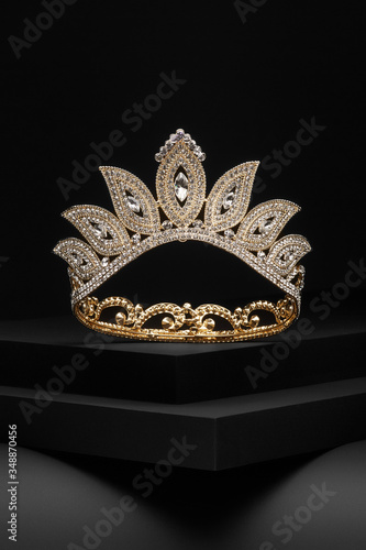 Subject shot of a tiara made as golden leaves adorned with diamond-like gems and clear sparkling rhinestones. The luxury queen crown is fixed on the black stepped surface.