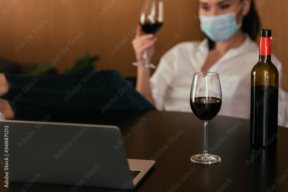 selective focus of woman in protective mask holding glass of red wine during video chat on laptop selective focus of woman in protective mask holding glass of red wine during video chat on laptop