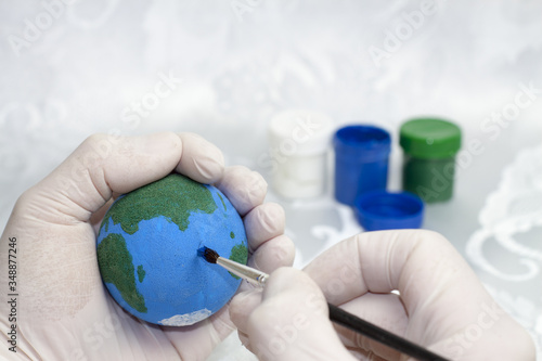 A human hand in white protective gloves painting the planet Earth on a styrofoam ball and in the background there are paints of blue, green and white.


