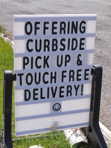 A business sidewalk sign regarding reopening during the Covid-19 outbreak says Offering Curbside Pick Up and Touch Free Delivery and has a smiley face to reassure customers during the Spring of 2020