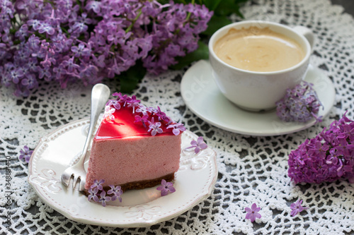 A berry mousse cake with chocolate base and juice jelly, decorated with lilac flowers, served with coffee.
