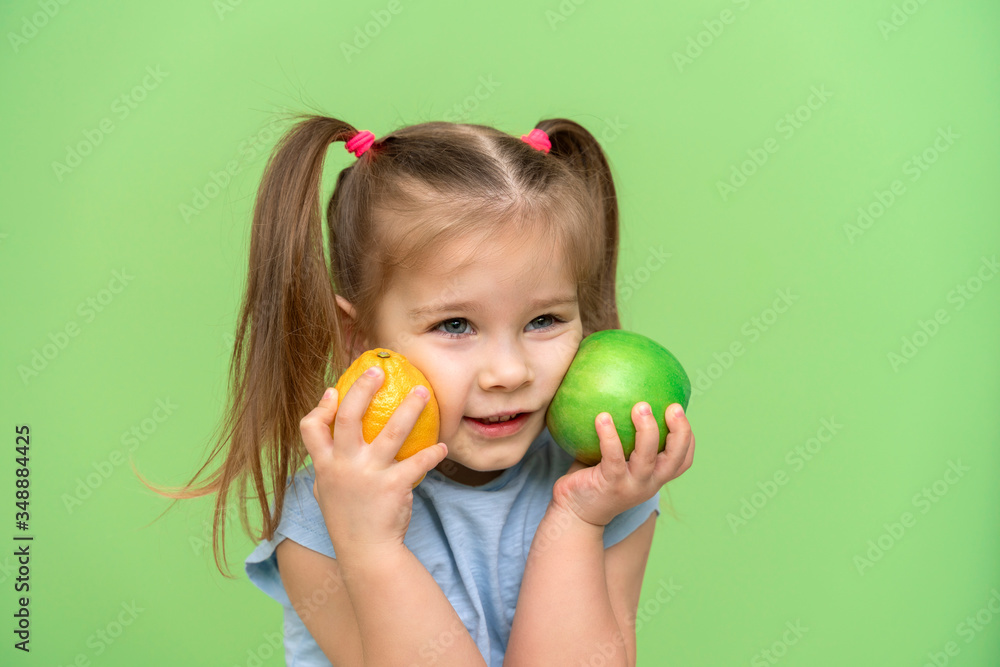 little girl 4 years old in a blue T-shirt on a green background put an apple and a lemon to her face and smiles. pre-school children healthy eating concept little girl 4 years old in a blue T-shirt on a green background put an apple and a lemon to her face and smiles. pre-school children healthy eating concept