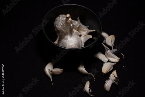 A bowl full of garlic cloves decorated with scattered pieces in a dark copy space background. Food and product photography.