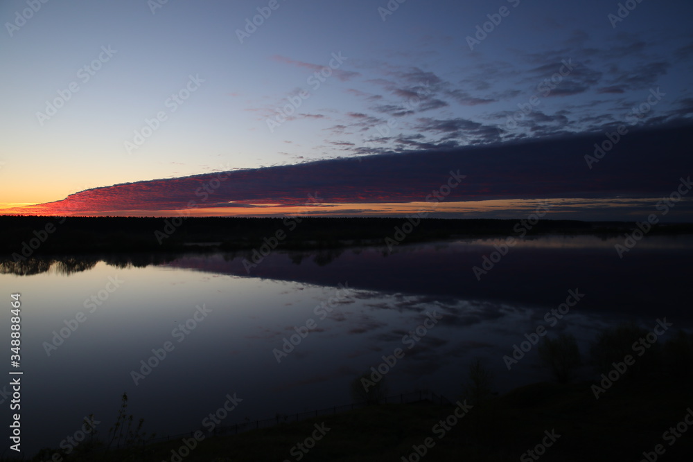 The water reflects the lilac sky the black forest by the river dividing the silvery surface from the clear horizon half covered with a mottled purple cloud in the mist at dawn.Landscape.Russia